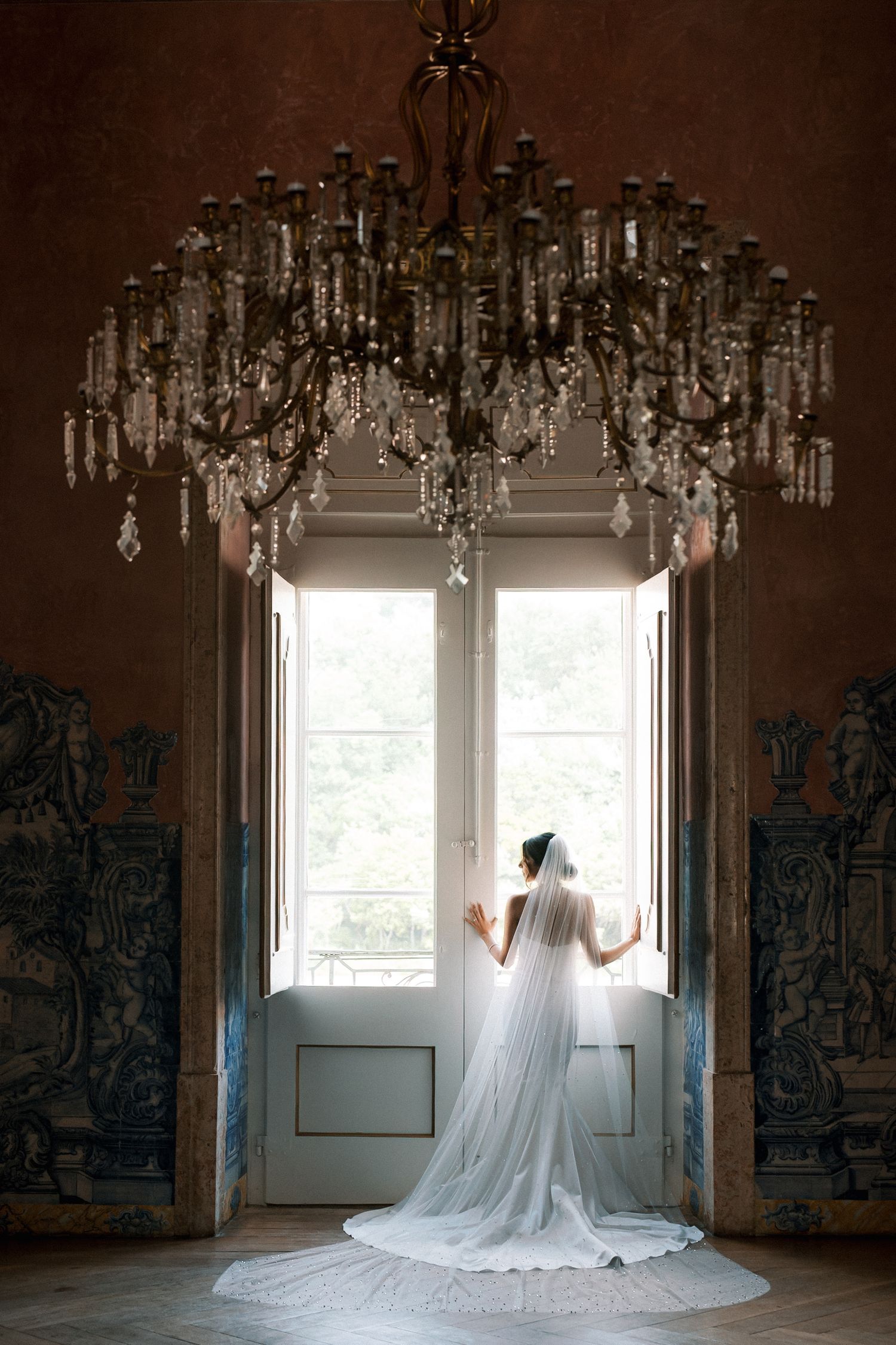 Bride portrait with long veil looking to a big window and a big chandelier on top at Palácio de Fronteira Lisbon palace room