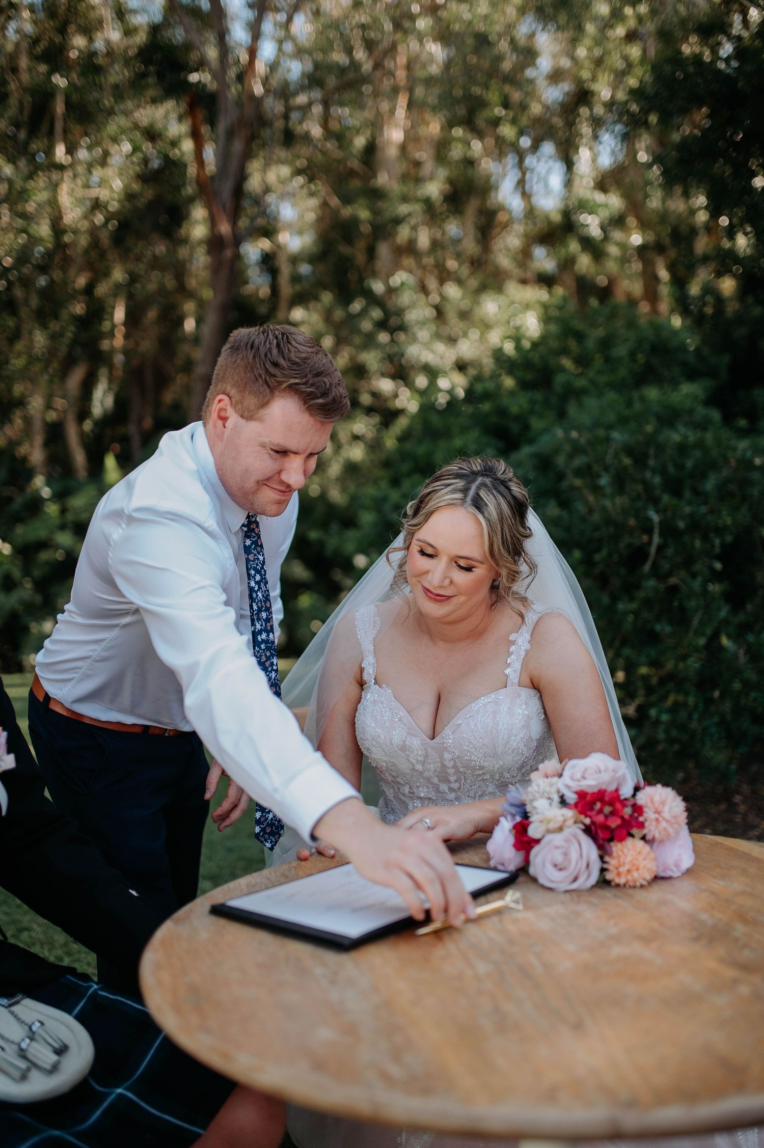 A couple in wedding attire signs documents together at a wooden table in an outdoor garden setting.