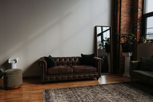 A brown leather Chesterfield sofa sits in a moody industrial loft space with wooden floors and exposed brick walls.