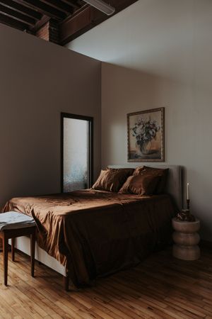 A moody bedroom with brown silk bedding, hardwood floors, exposed beams, and framed artwork on gray walls.