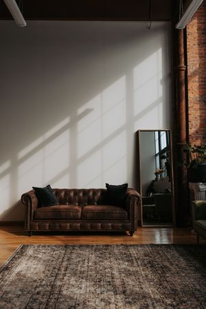 A brown leather tufted sofa sits against exposed brick walls in a minimalist room with dramatic window lighting.
