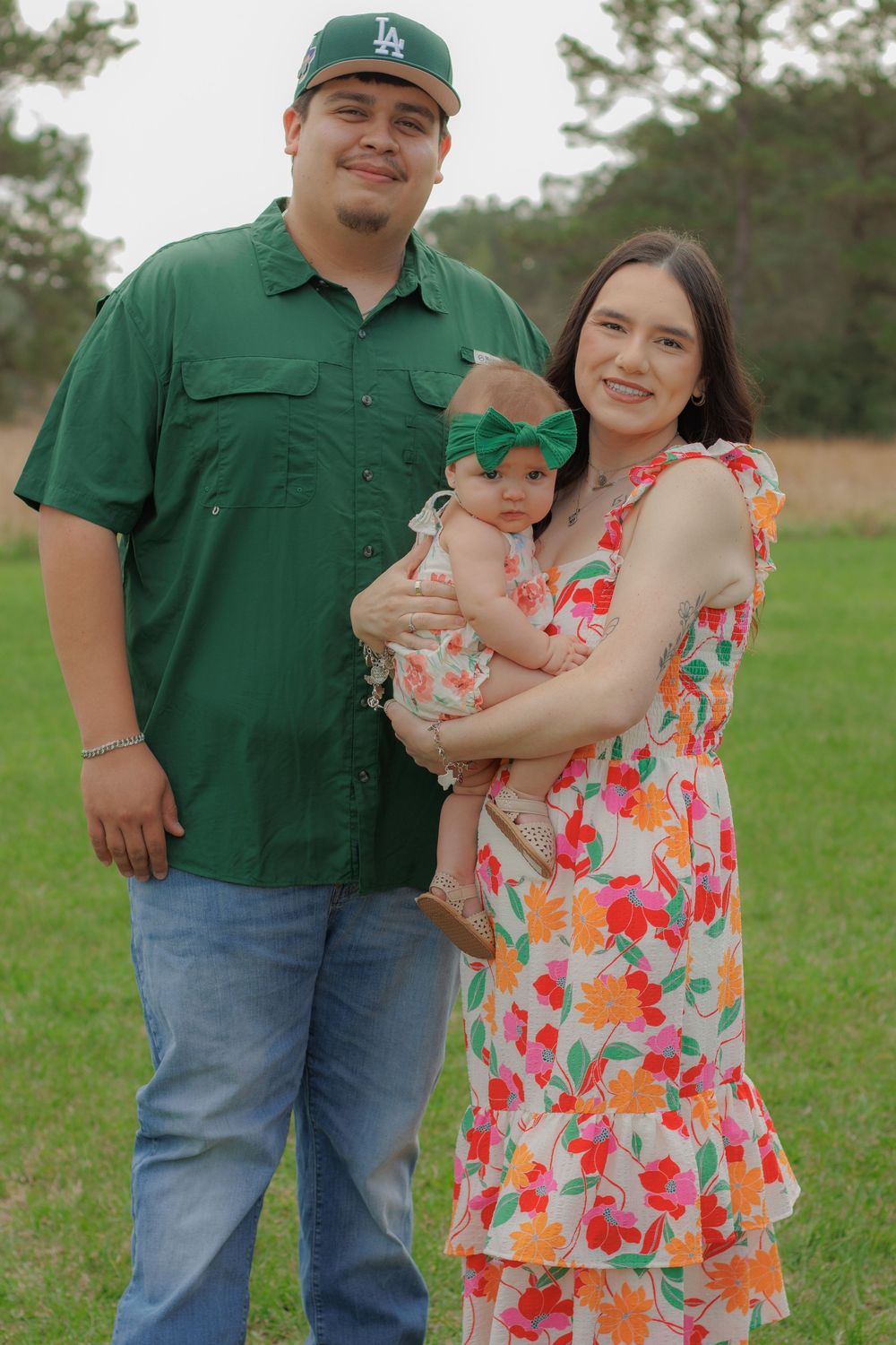 A happy family of three, a man wearing a green shirt, a woman wearing a floral dress, and a young child, posing together in a grassy field.