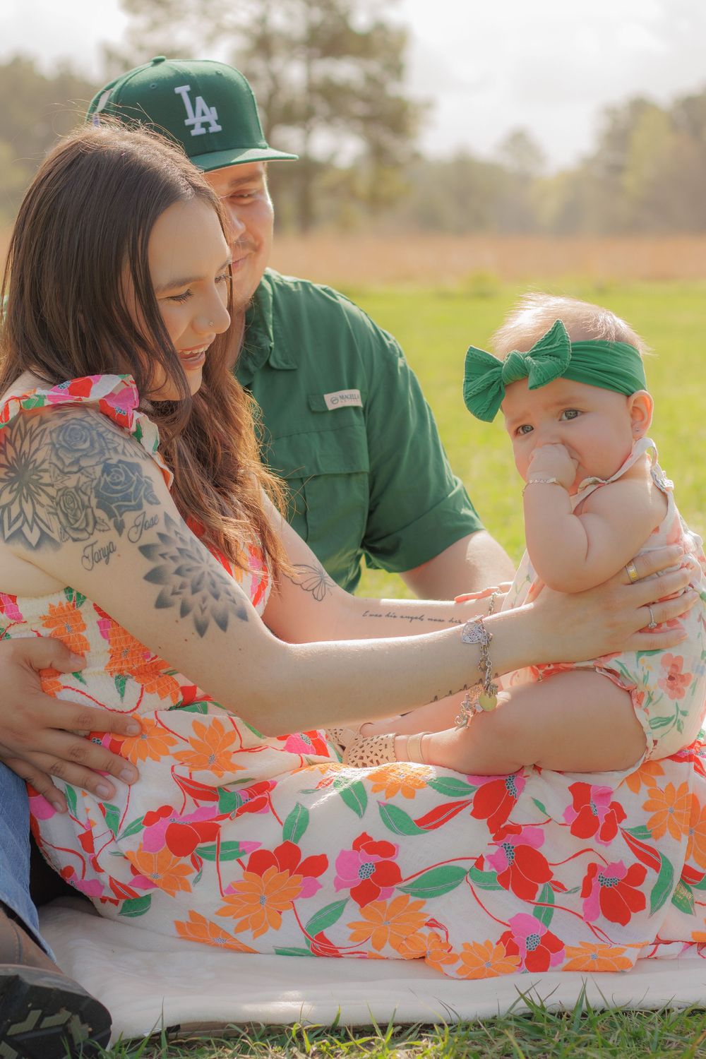 A woman wearing a floral dress holds a baby while a man in a green shirt stands next to them in a grassy field.