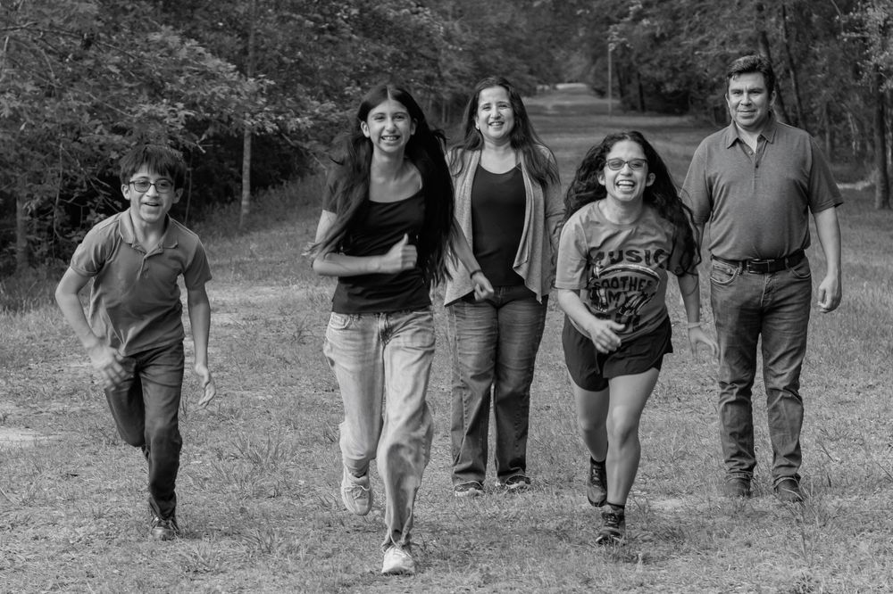 Black and white photo of a family of five running and walking through a grassy field near trees.