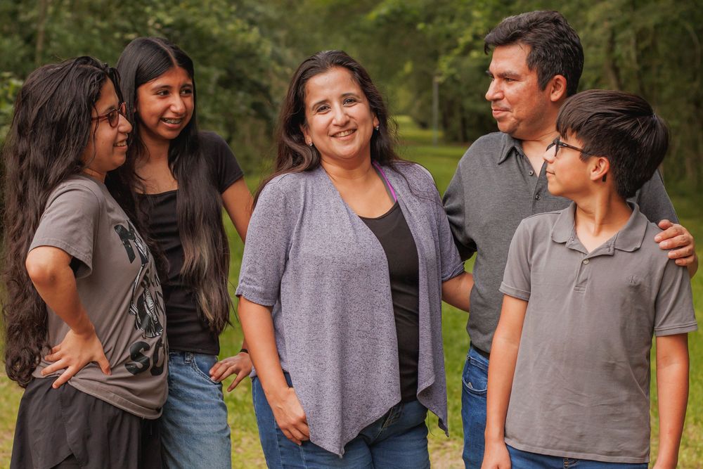 A happy Hispanic family of five smiling and talking together outdoors in a lush green park setting.