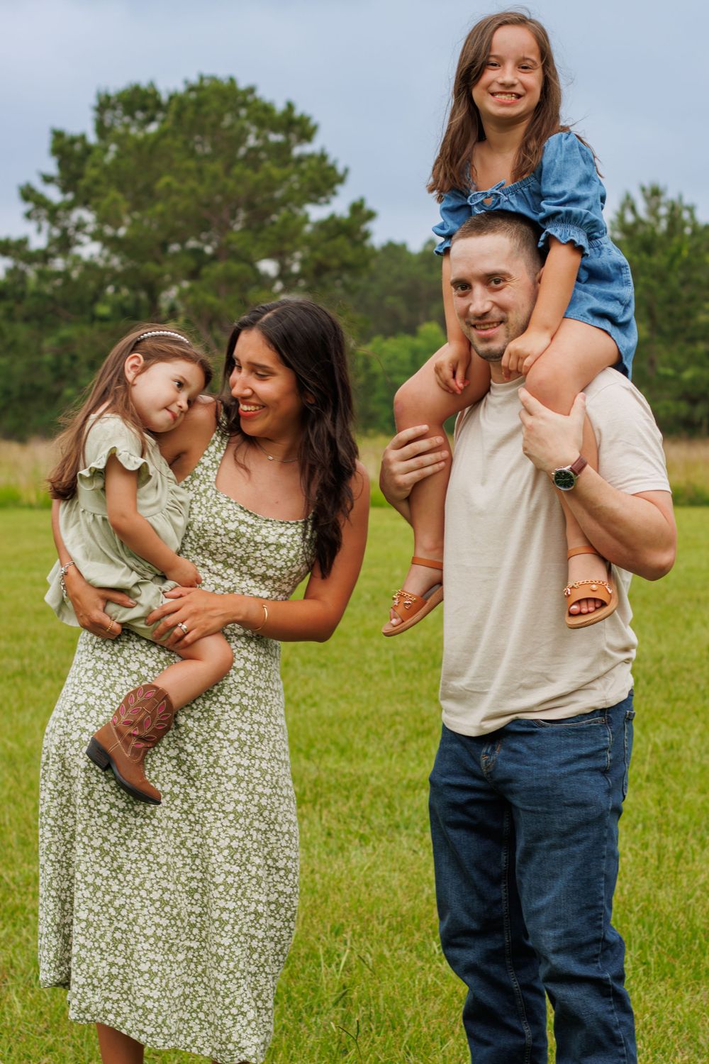 Happy family of four posing outdoors in a green field, mother holding toddler, father with girl on shoulders.