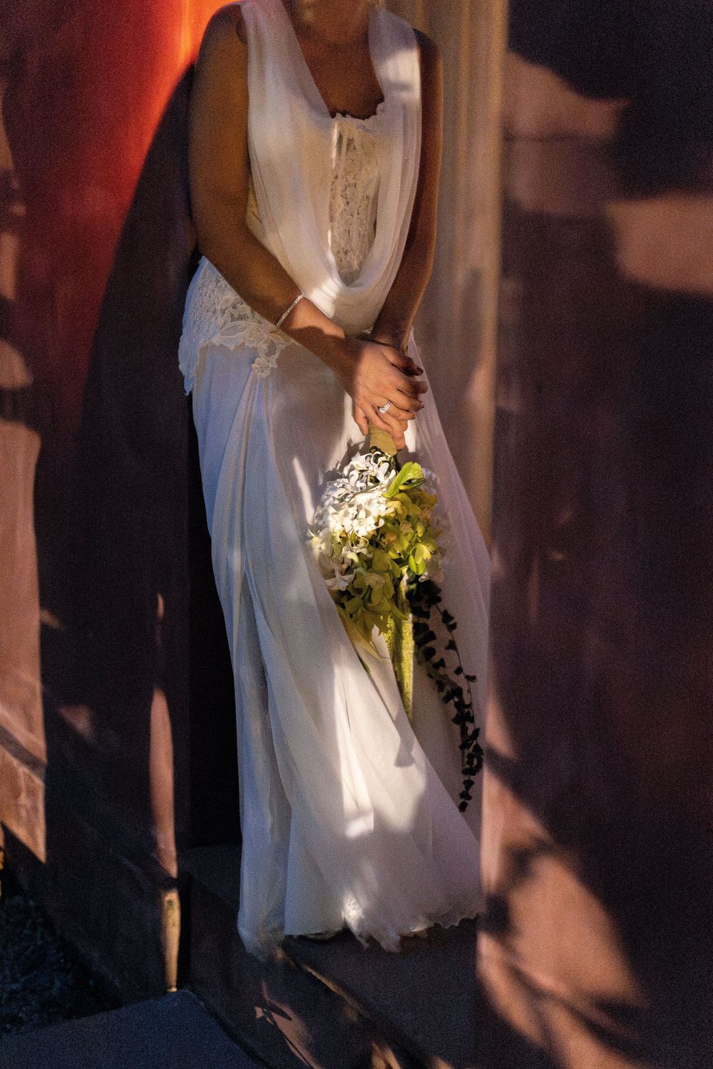 A bride in a white lace wedding dress holds a cascading bouquet of white and green flowers in dramatic lighting.