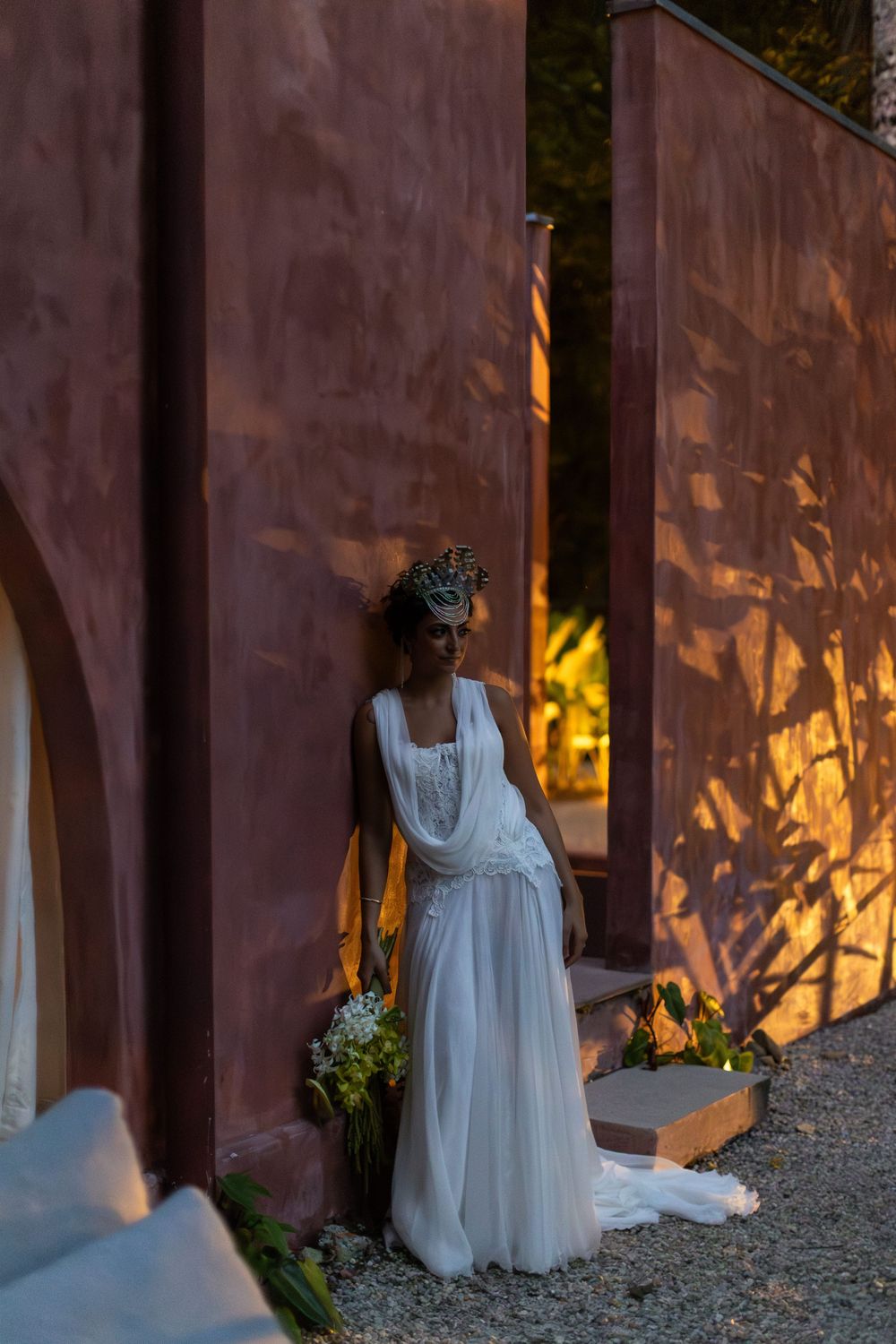 A bride in a flowing white dress stands against a rustic wall with dramatic sunset lighting creating shadow patterns.