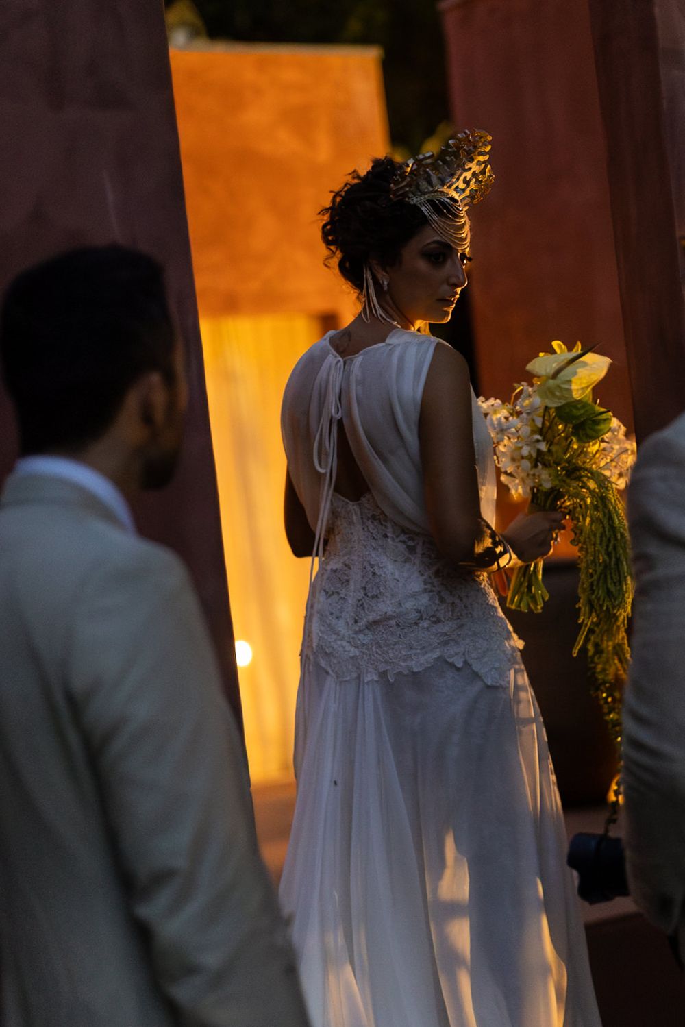A bride in a white lace dress holds a sunflower bouquet while standing in a warmly lit indoor venue.