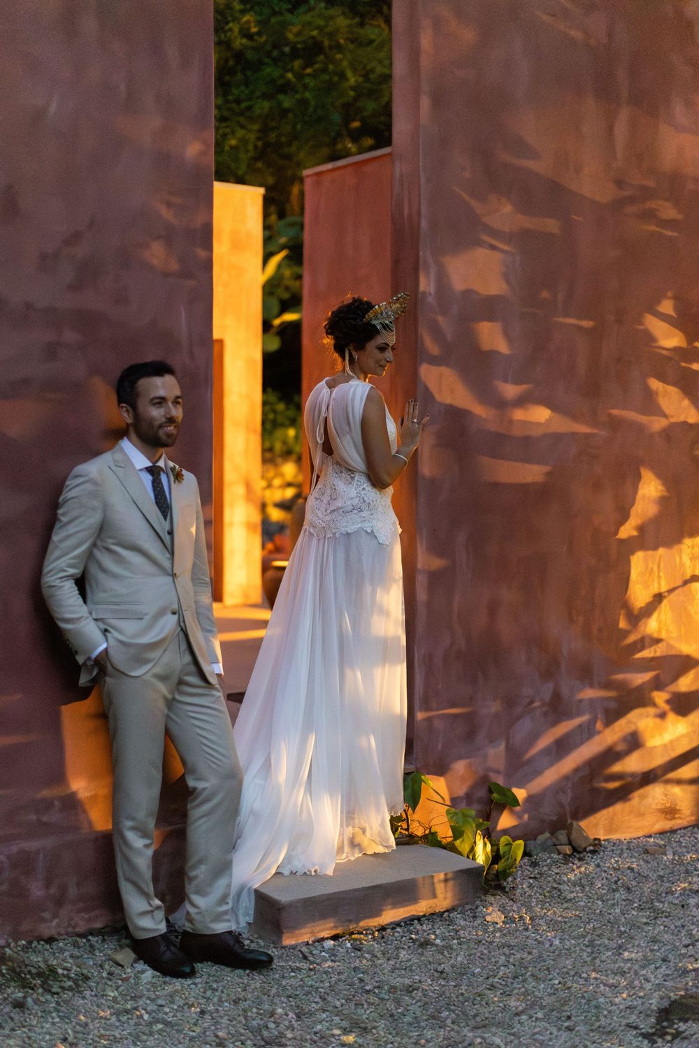 A couple in wedding attire poses against a rustic terracotta wall with dramatic sunset lighting creating shadow patterns.