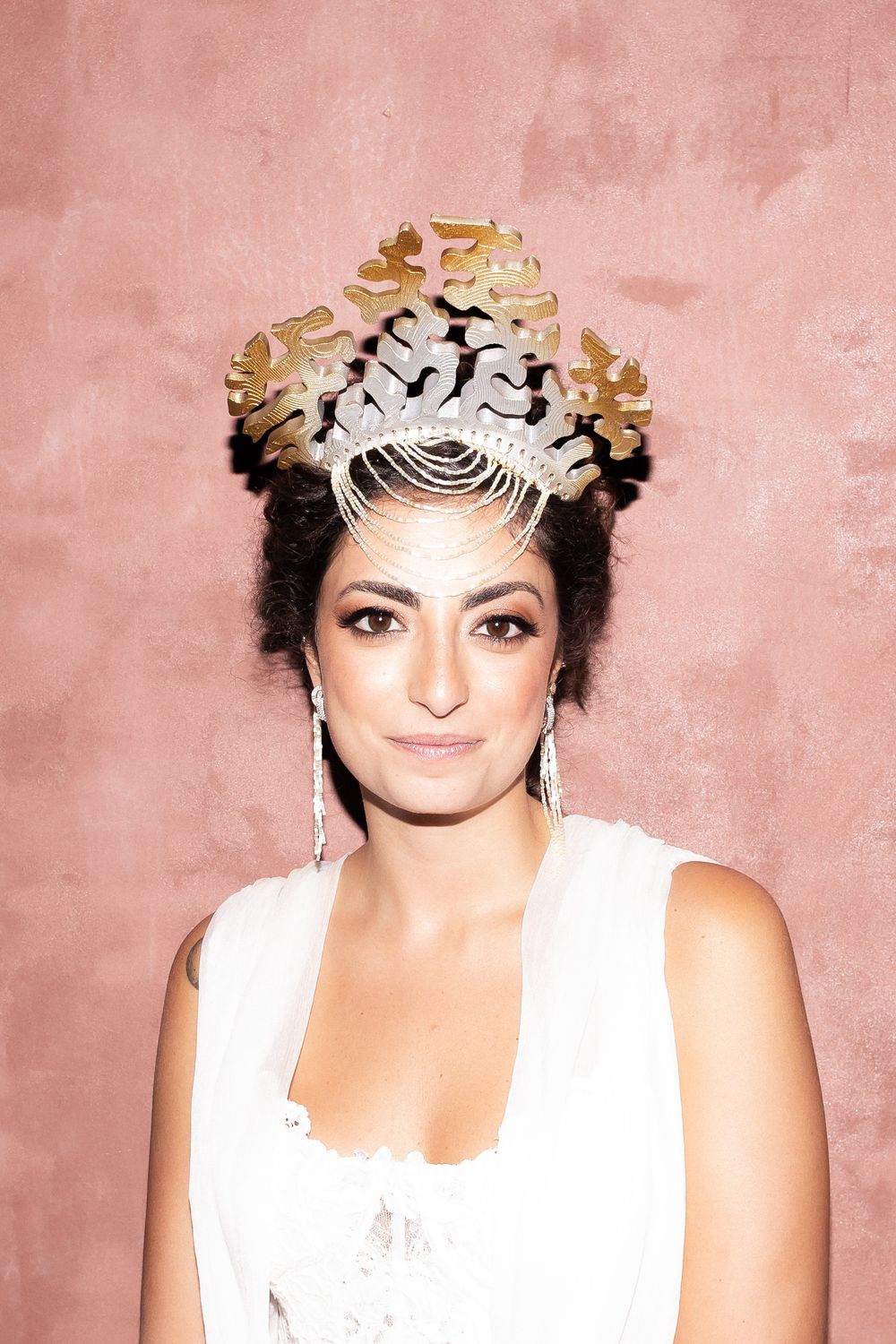 A person wearing a white sleeveless top and gold floral headpiece poses against a pink background.