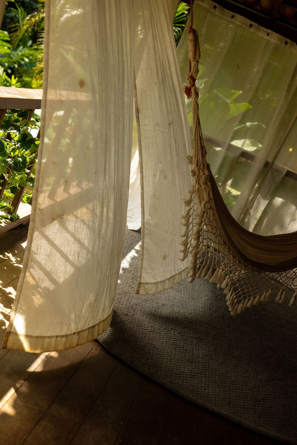 Sunlight filters through sheer white curtains creating soft shadows on a woven hammock in a peaceful corner.