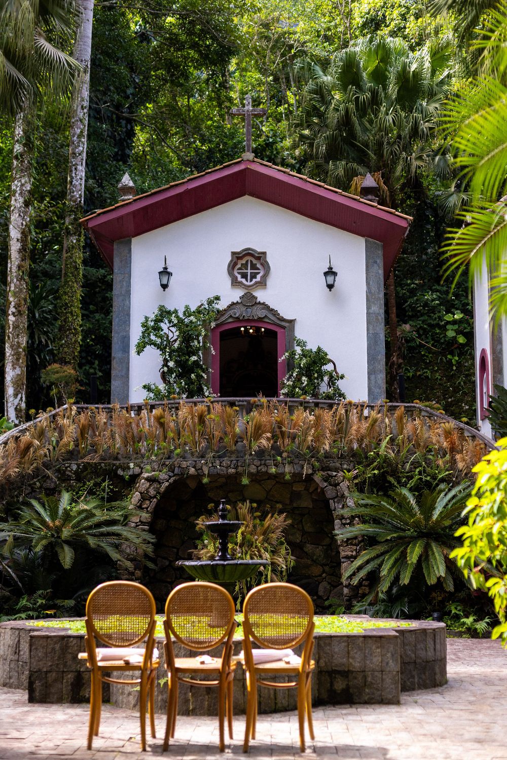 Three wooden chairs sit in front of a stone arch walkway leading to white chapel buildings in a tropical garden setting.