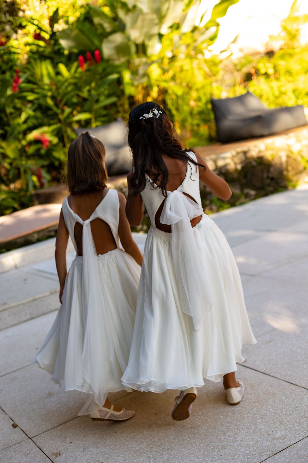 Two flower girls in matching white dresses with bow details walk together on a sunlit garden path.