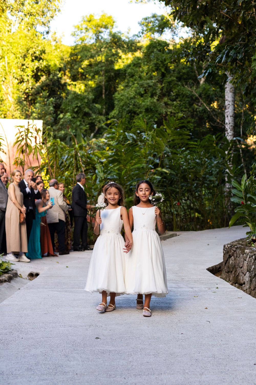 Young flower girls in white dresses walk down a light-colored pathway at outdoor wedding.