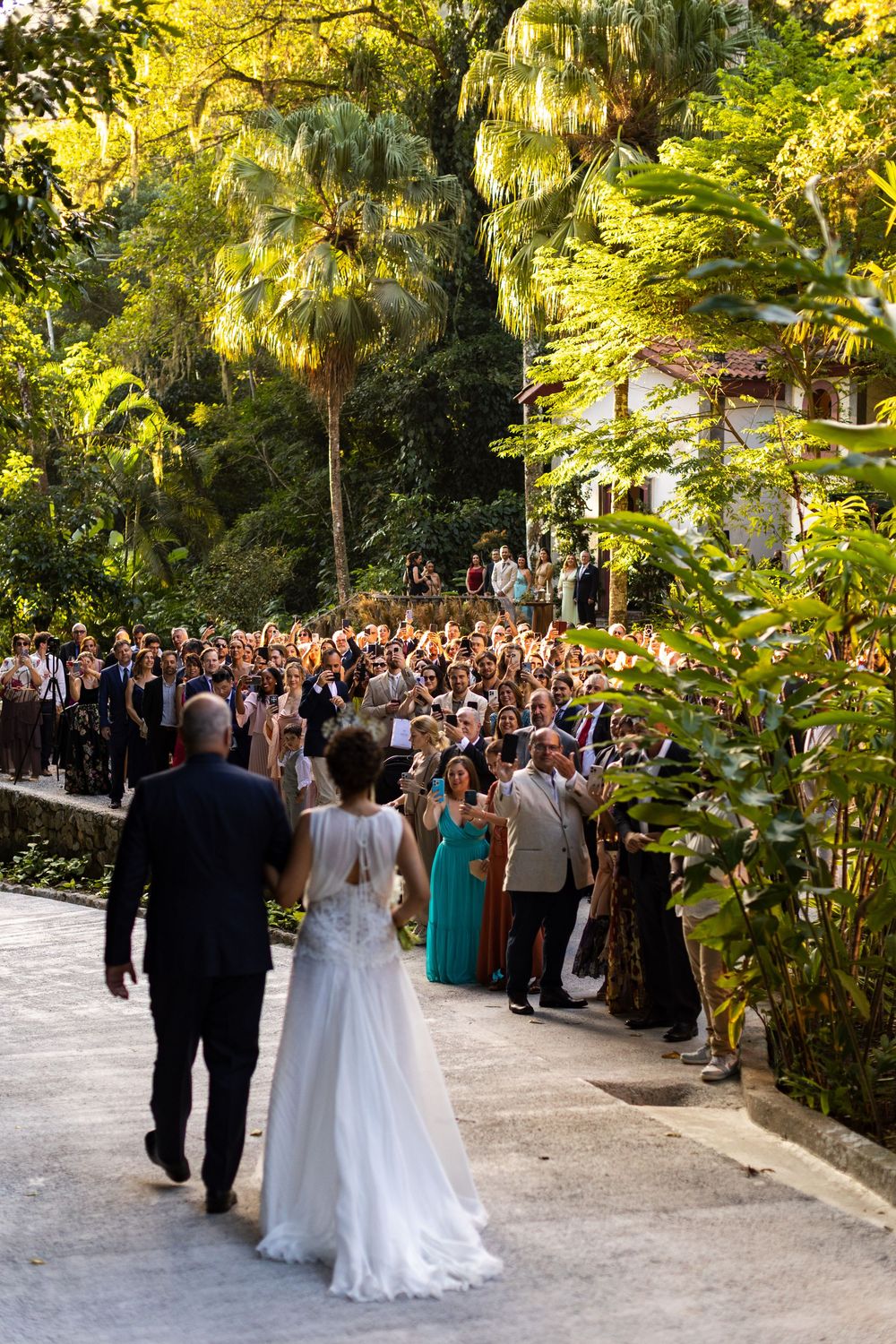 Wedding couple walks down garden path surrounded by large crowd of guests at sunset ceremony.