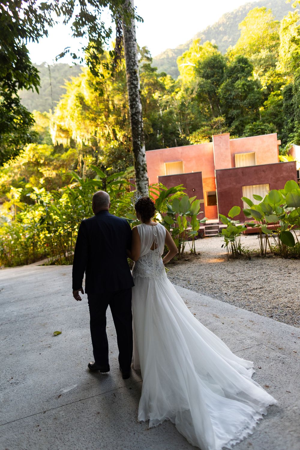 A couple walks down a path with pink buildings and lush greenery in the background.
