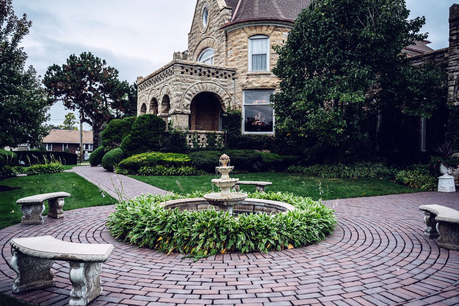 Circular brick patio with ornate fountain and stone benches in front of Victorian mansion with landscaped gardens.