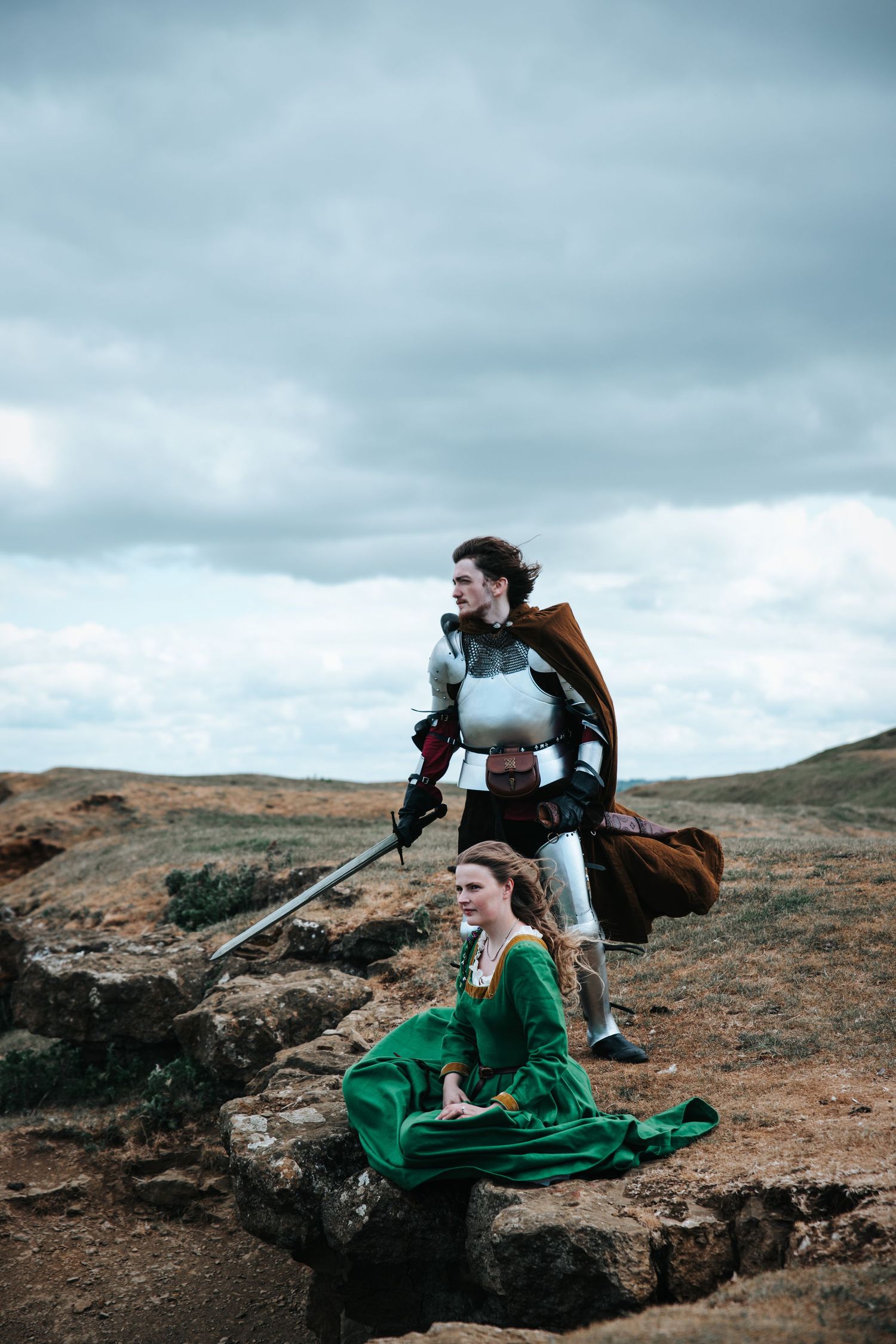 Adventurous figures in medieval-inspired costumes explore a rugged mountainous landscape under moody cloudy skies.