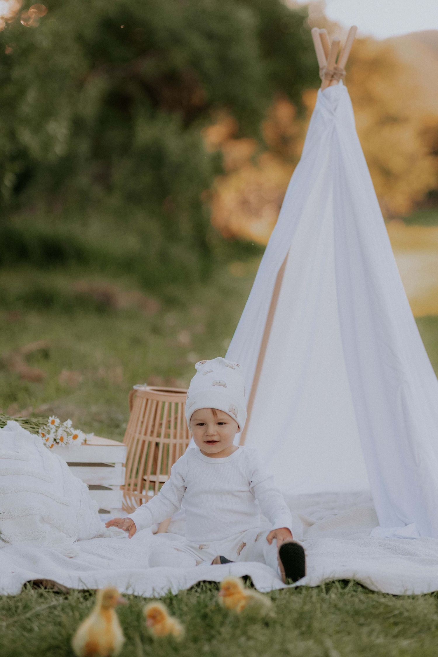 Bábätko v bielom oblečení sedí pri teepee stane v obklopení malých žltých káčat. Rozprávkové detské fotenie v prírode, Žilina.