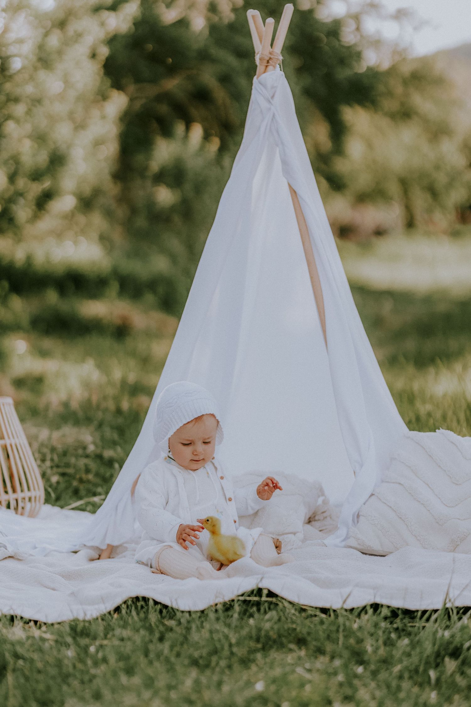 Bábätko v bielom čepčeku sedí pred teepee stanom v záhrade so žltými káčatkami. Rozprávkové jarné fotenie detí v Žiline.