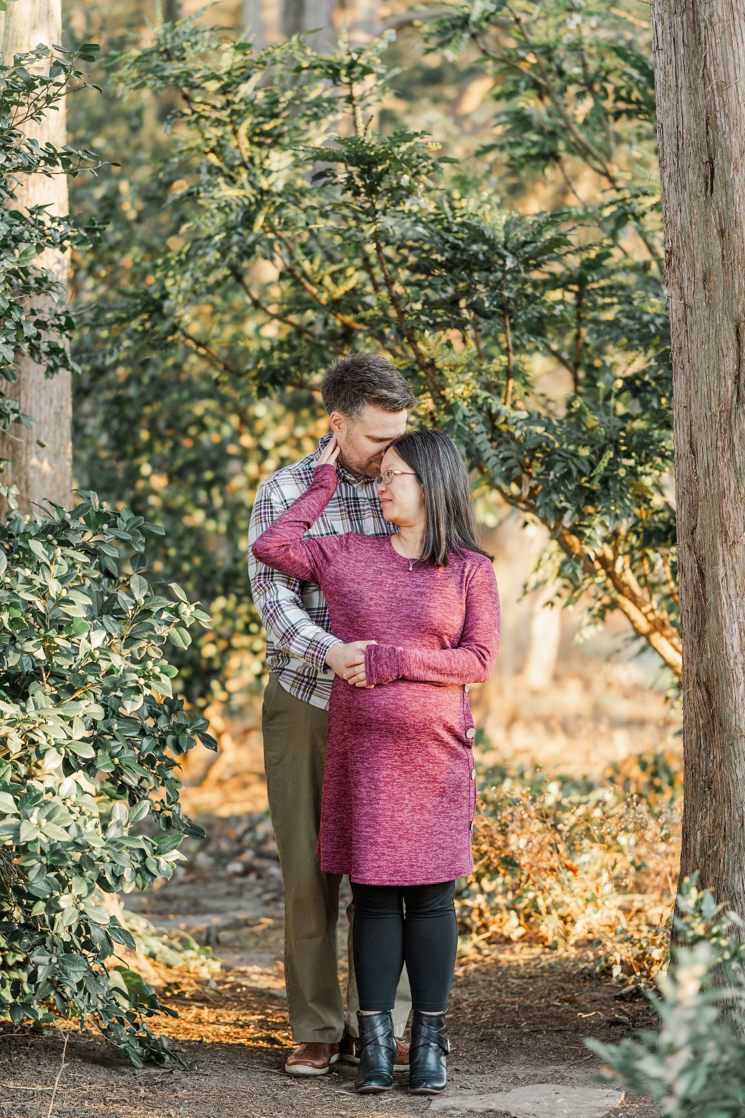 A couple shares a tender moment among autumn trees and fallen leaves in a park setting.