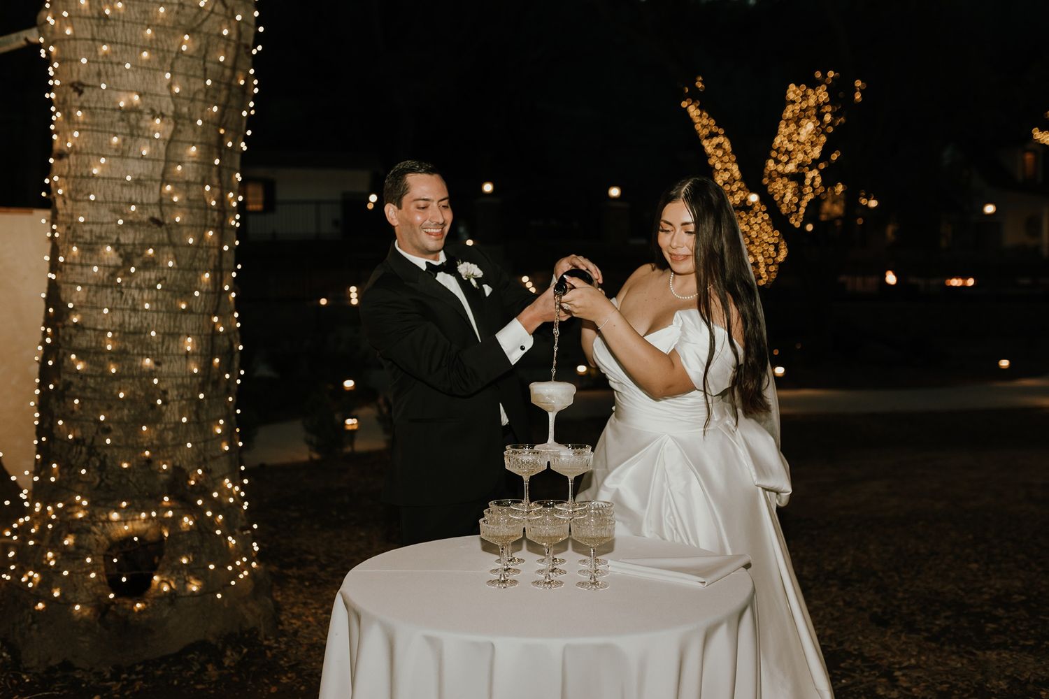 Wedding couple pours champagne at an evening reception with twinkling lights in the background at The Oaks at Duncan Lane.