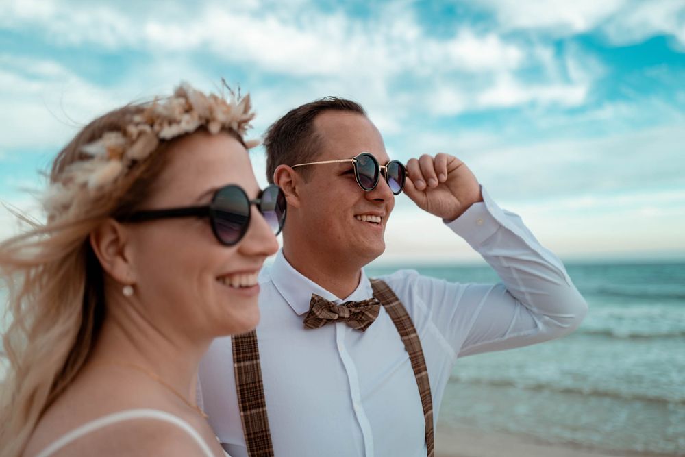Lachendes Brautpaar mit Sonnenbrillen am Strand. Entspannte und fröhliche Atmosphäre ohne steife Posen