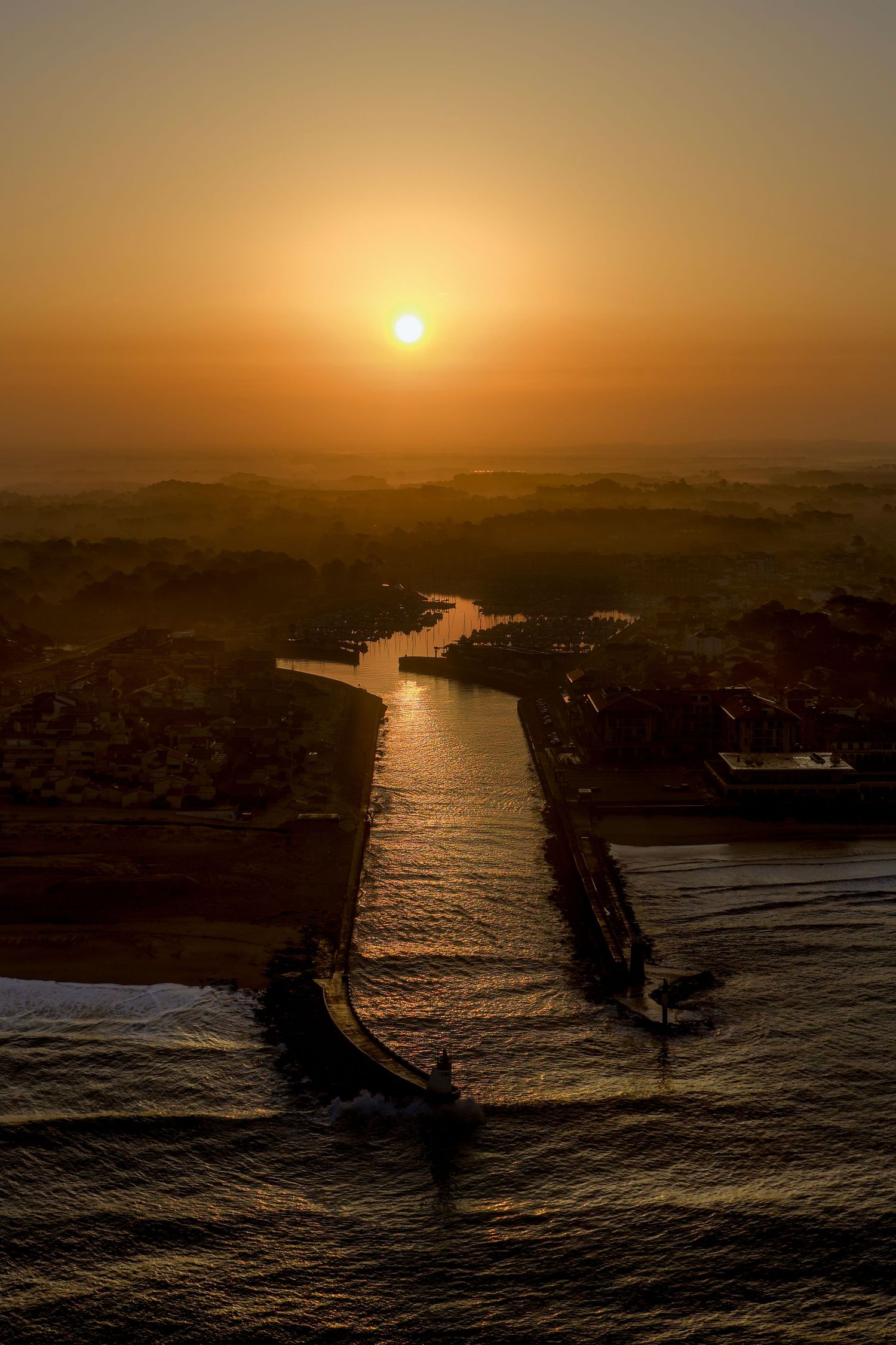 Aerial view of a winding river reflecting golden sunlight during sunset over rugged coastal cliffs and ocean waves.