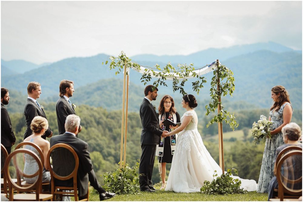A mountain wedding ceremony takes place under a rustic wooden arch decorated with greenery and flowers.