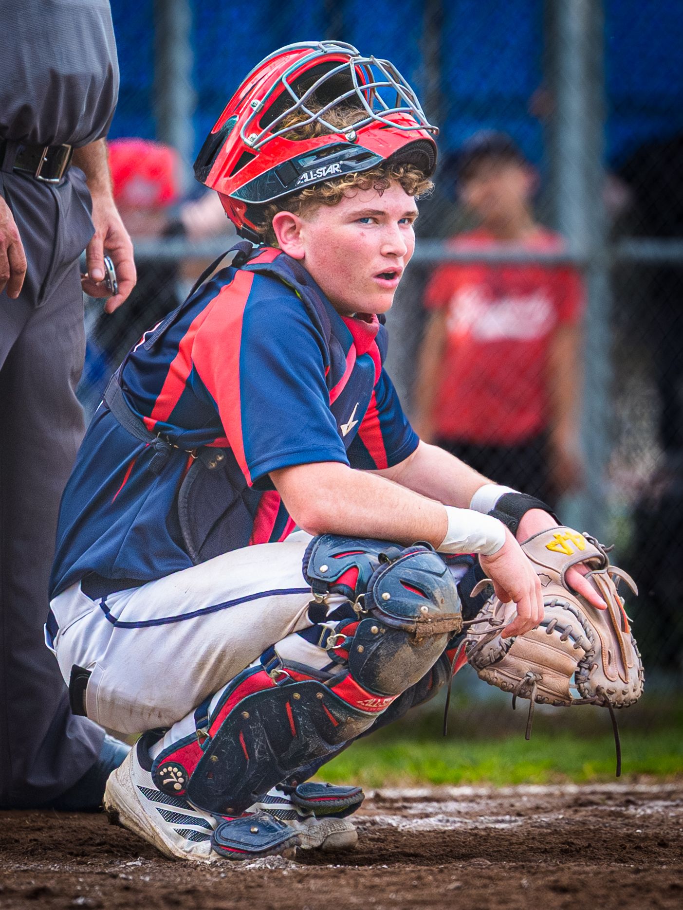 Baseball catcher in blue and red uniform crouches low to block a play at home plate during a game.