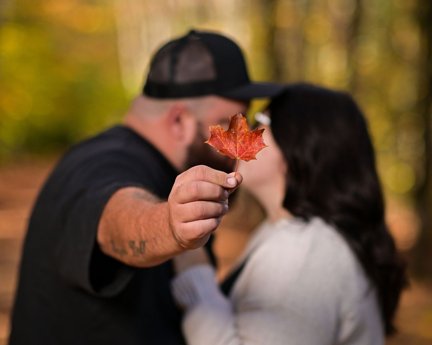 A bright red autumn maple leaf is held up in focus against a soft blurred forest background.