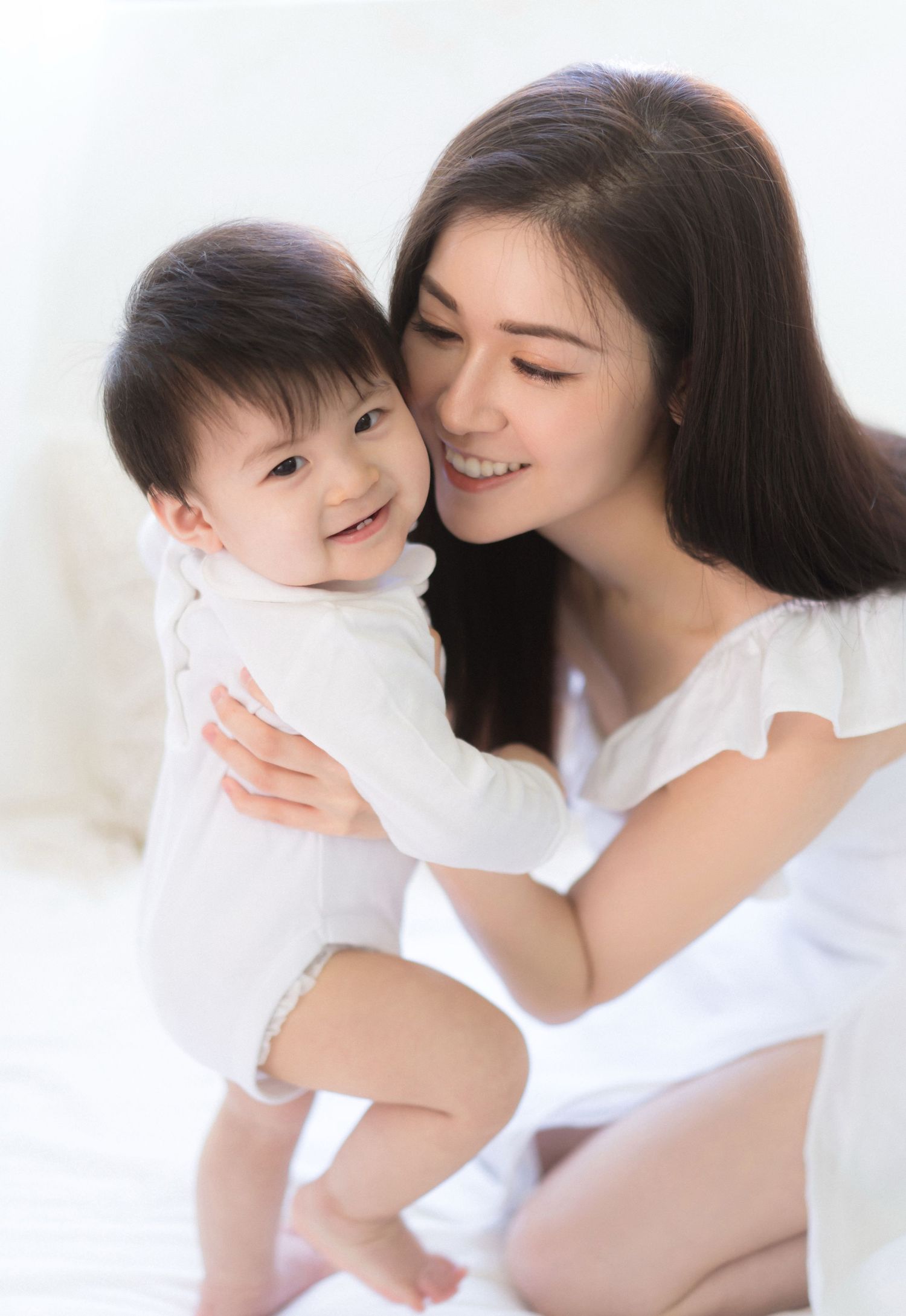 A smiling young Asian woman in white holds a happy baby in a white onesie on a bright, white background一位面帶笑容的年輕亞裔女子穿著白色衣服，抱著一個穿著白色連身衣的快樂嬰兒，
