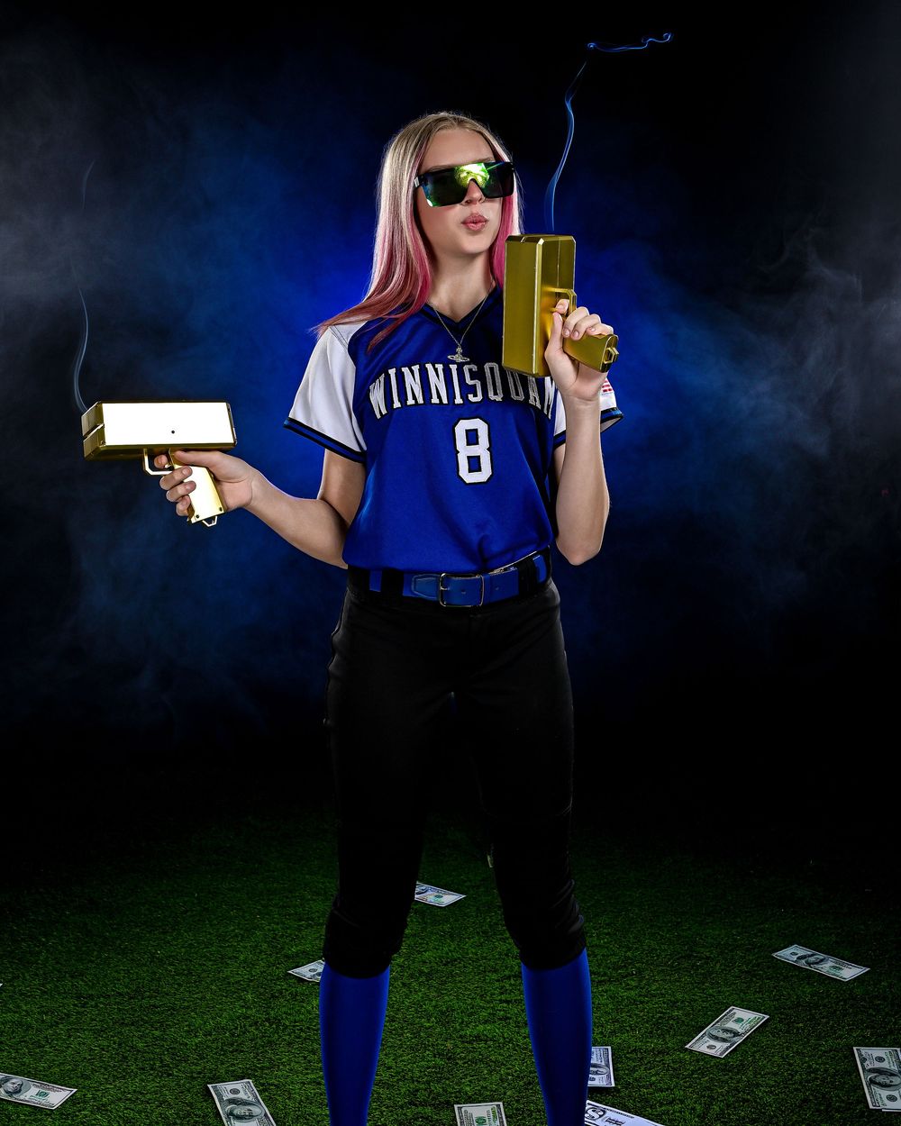 Softball player in blue Winnisquam jersey #8 posing with gold guns and sunglasses on turf with money.