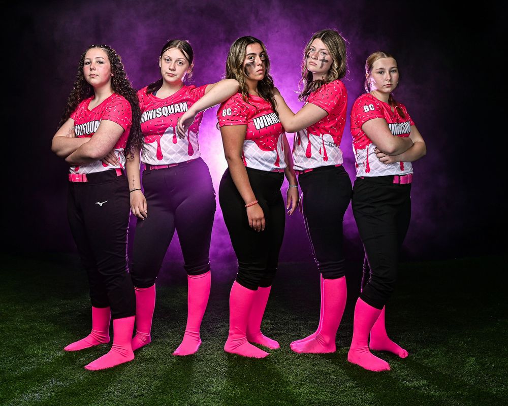 Five women in matching pink and black softball uniforms pose confidently on a turf field with dramatic purple smoke background.