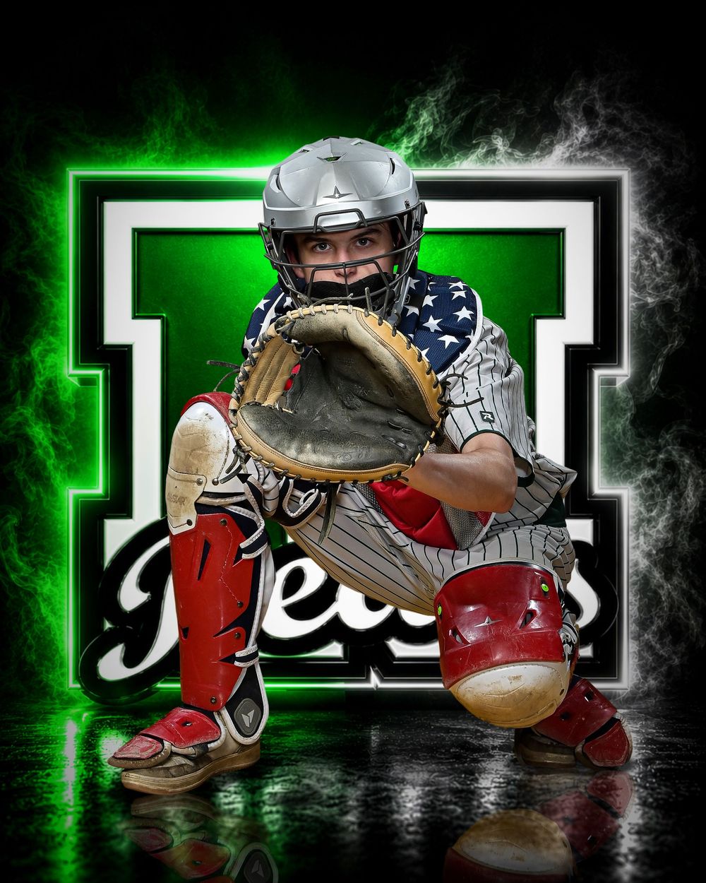Youth baseball catcher in full gear with American flag jersey, crouching in stance before green team logo backdrop.