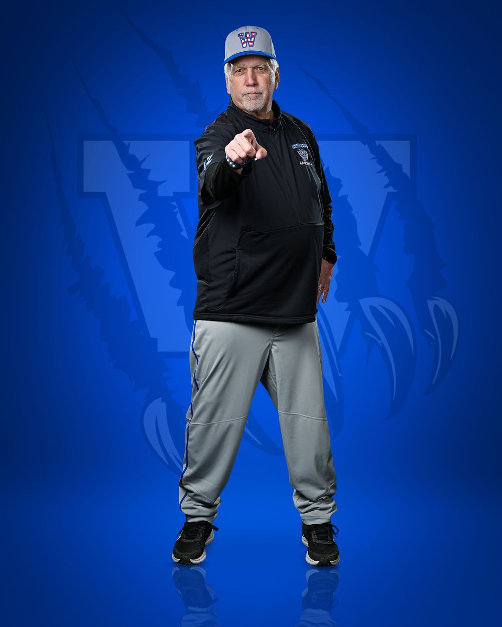 Coach in black jacket and gray pants points forward against a blue background with a large W logo.
