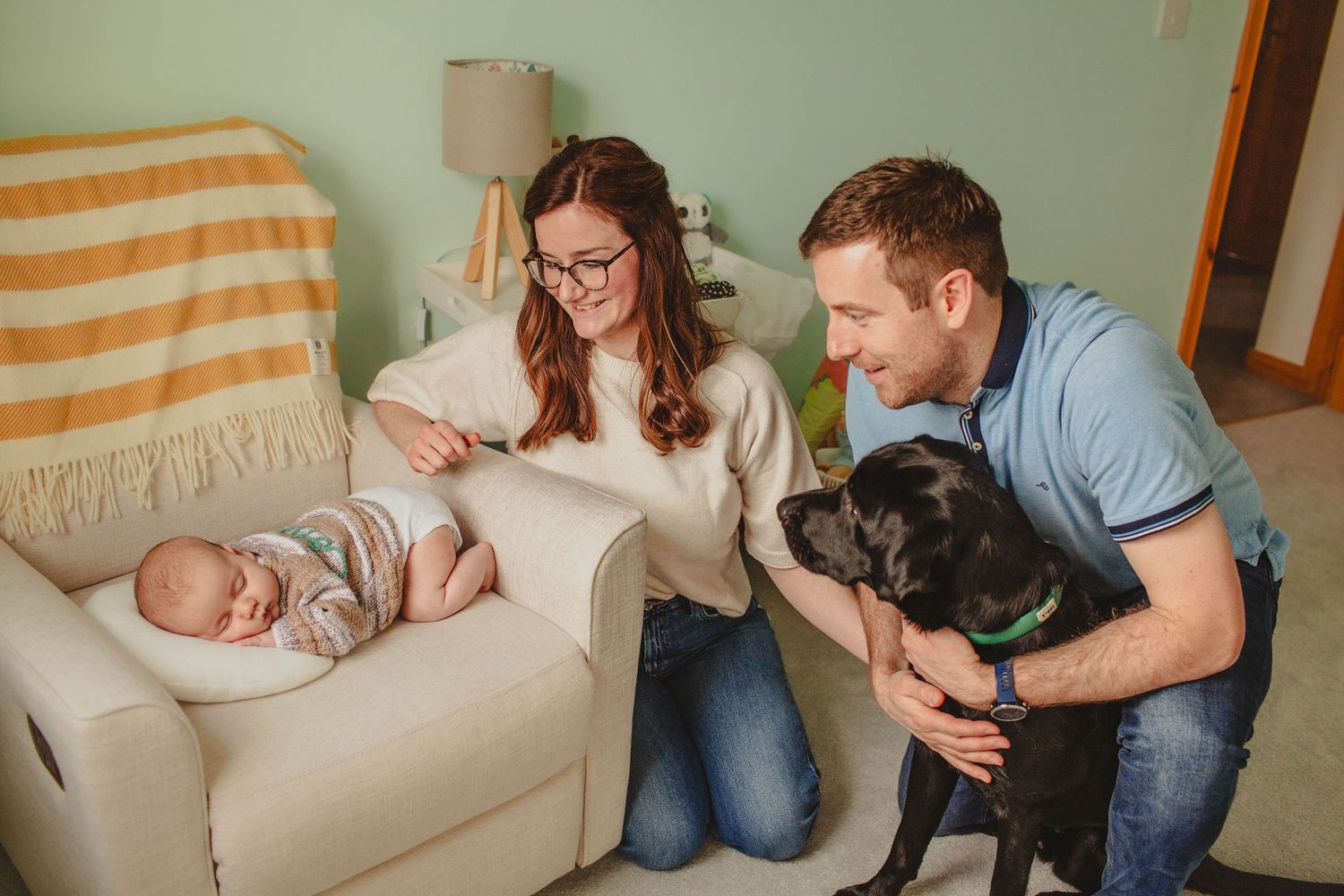 A couple sits with their sleeping baby on a couch while their black dog joins them in a cozy living room setting.
