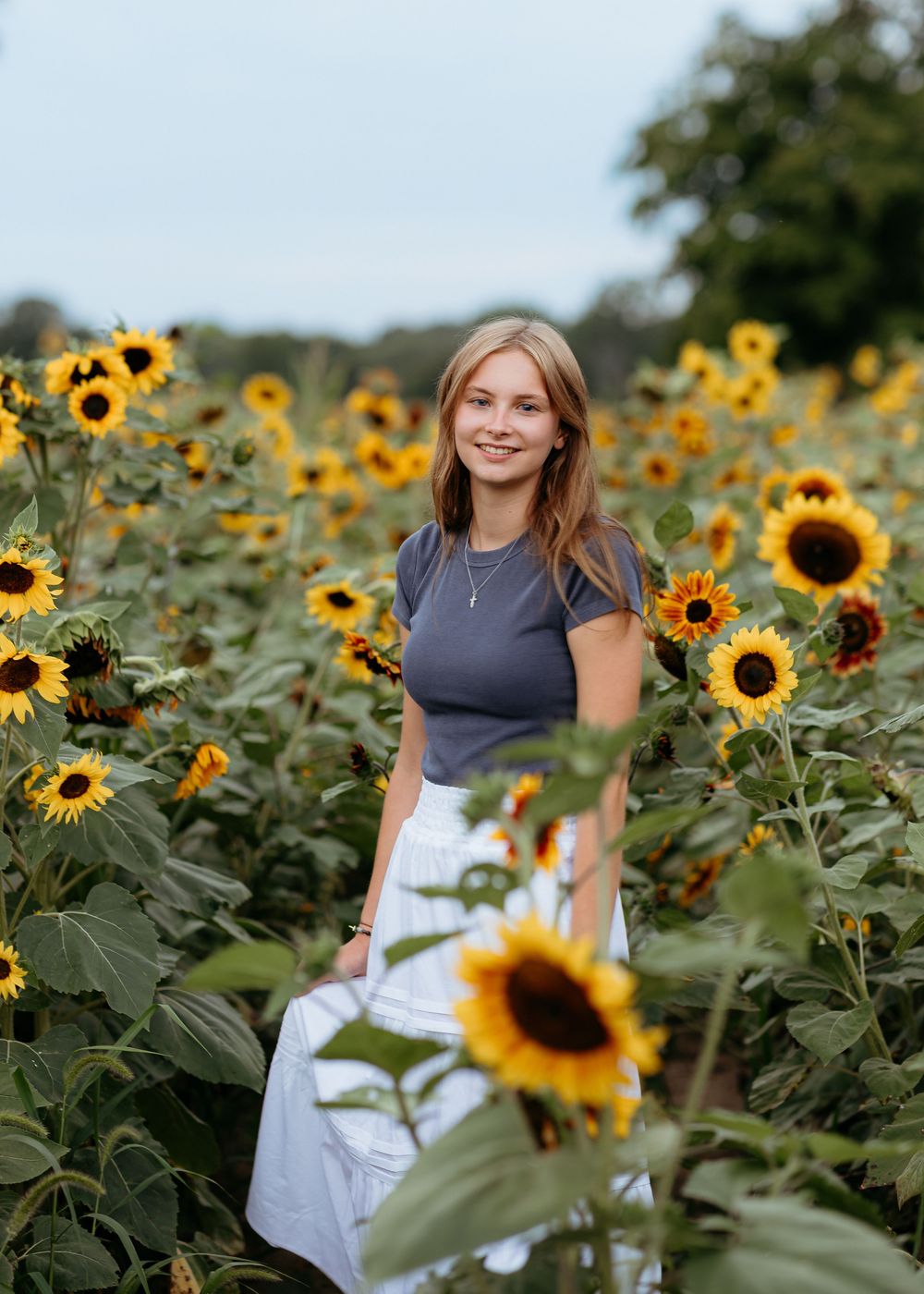 Girl in white dress poses in vibrant sunflower field on sunny day. Sunflower photo sessions. Senior photographer. Central Michigan.