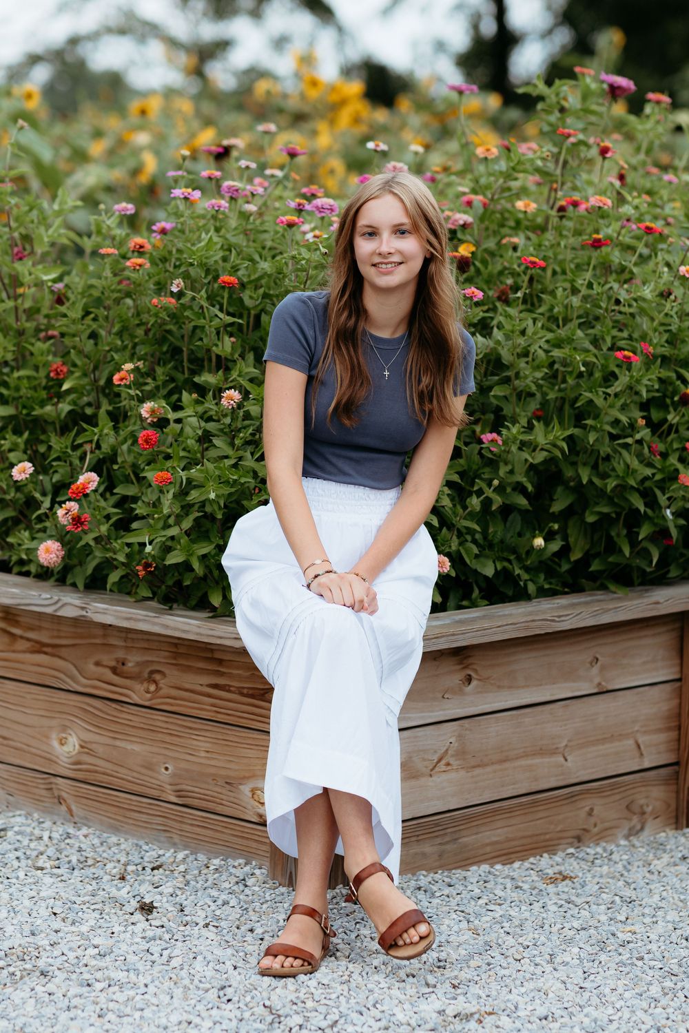 Girl in cute summer casual attire sits on wooden planter box surrounded by colorful flowers. Senior photos with wildflowers central Michigan