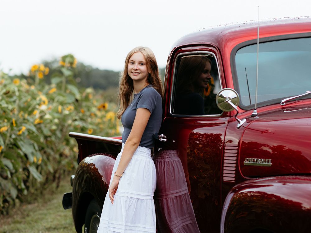 Senior girl in white dress stands next to vintage red pickup truck in sunflower field. Truck senior photos. Central Michigan senior photos