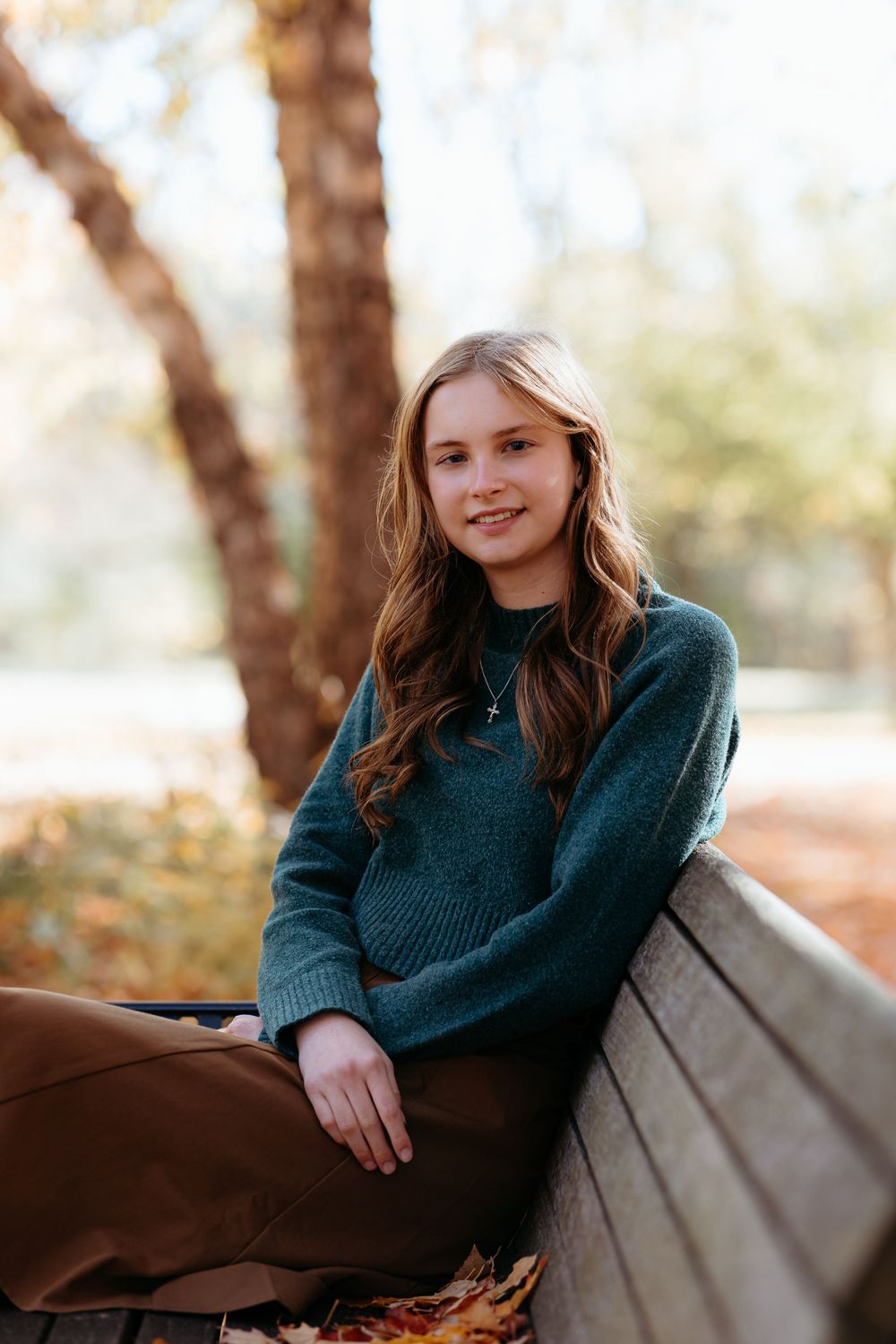Young person in a dark teal sweater poses on a wooden bench in an autumn outdoor setting.
