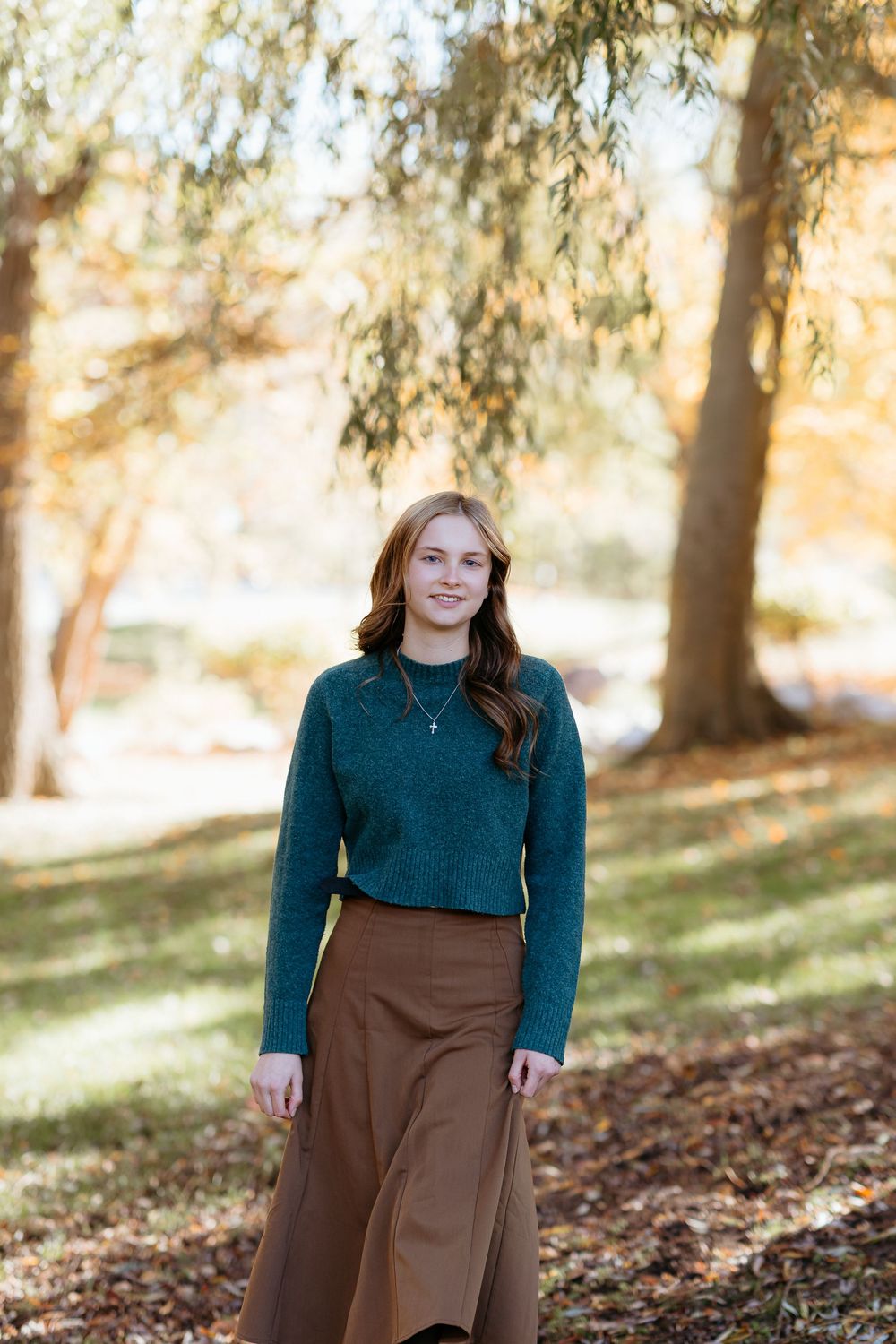 Person wearing teal sweater and brown skirt poses among fall trees on grassy slope.