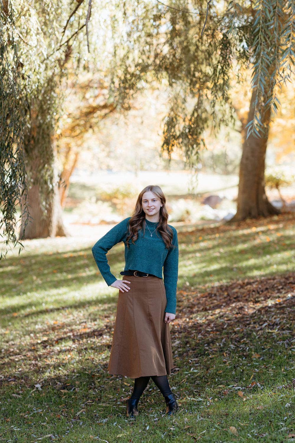 Girl in teal top and brown dress stands on leaf-covered ground with willow trees in background. Outdoor senior photos. Central Michigan.