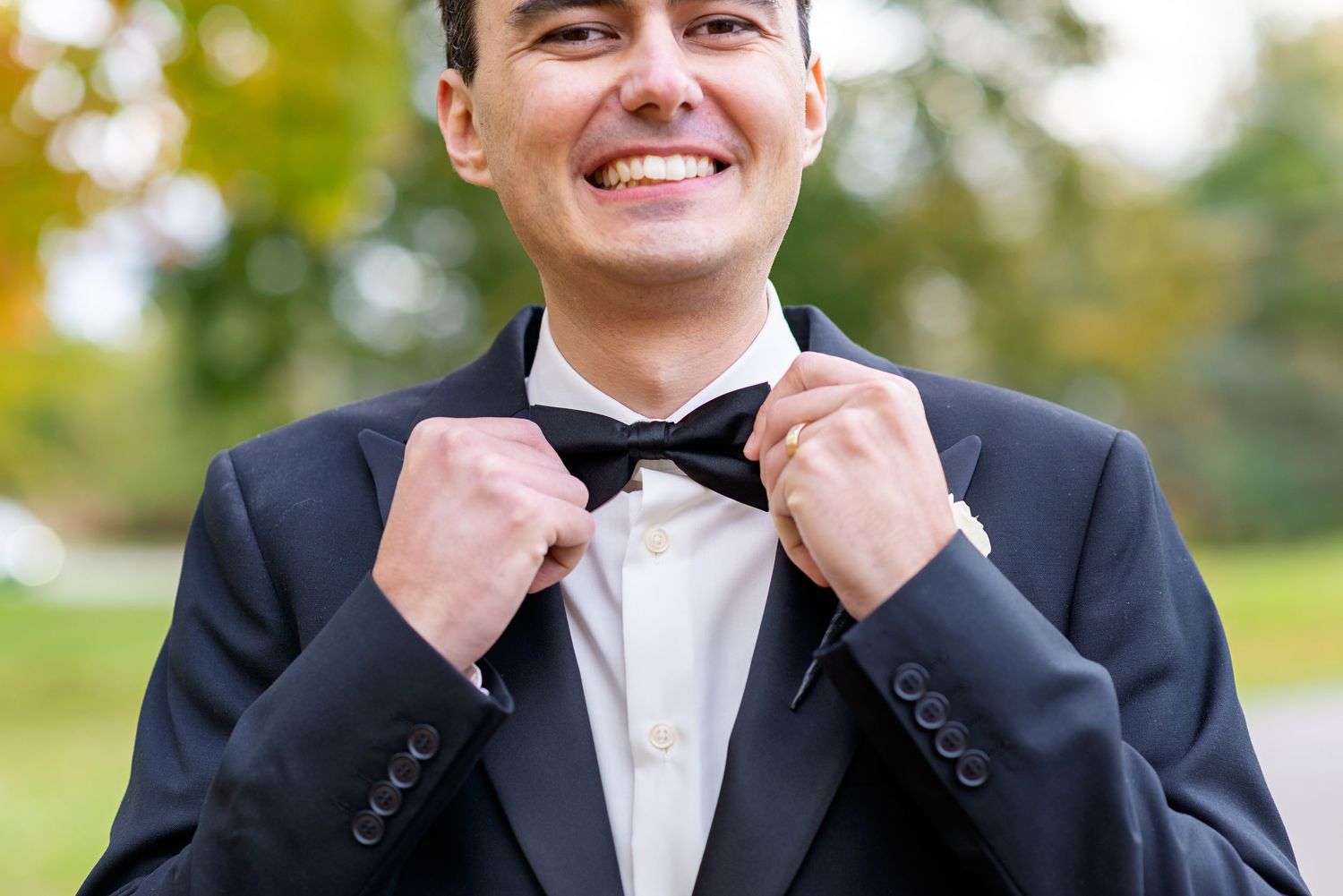 A person in a black tuxedo adjusts their bowtie while standing outdoors with a blurred natural background.