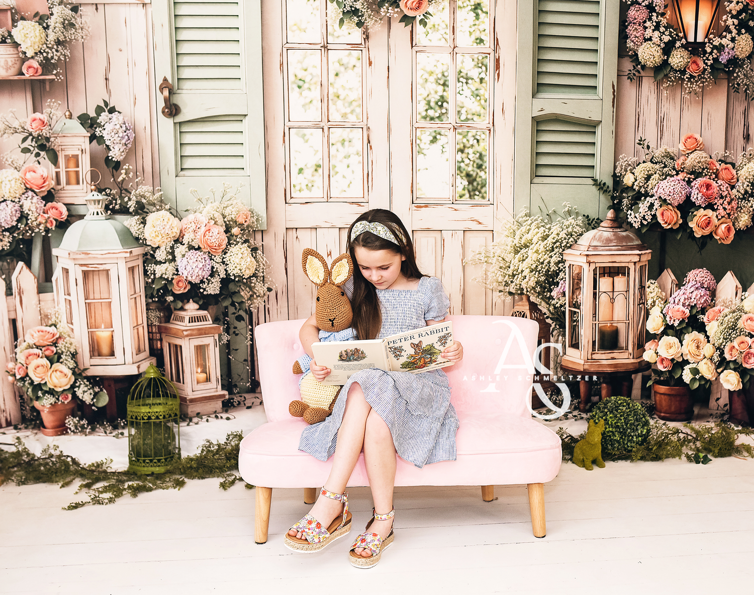 Young girl in blue dress reading book with stuffed bunny on pink sofa in floral spring studio setting.