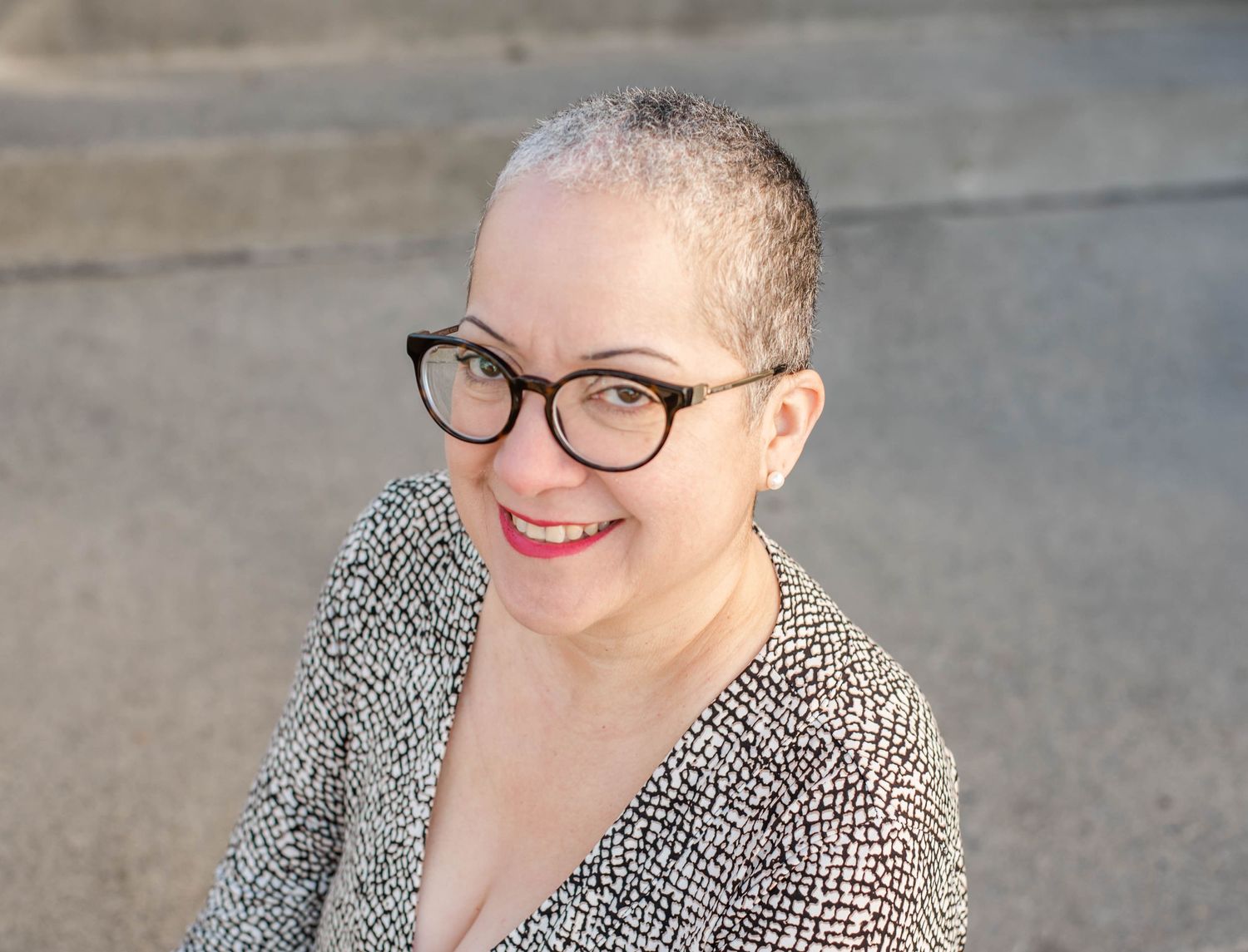 Woman in patterned dress and glasses stands smiling against urban backdrop.