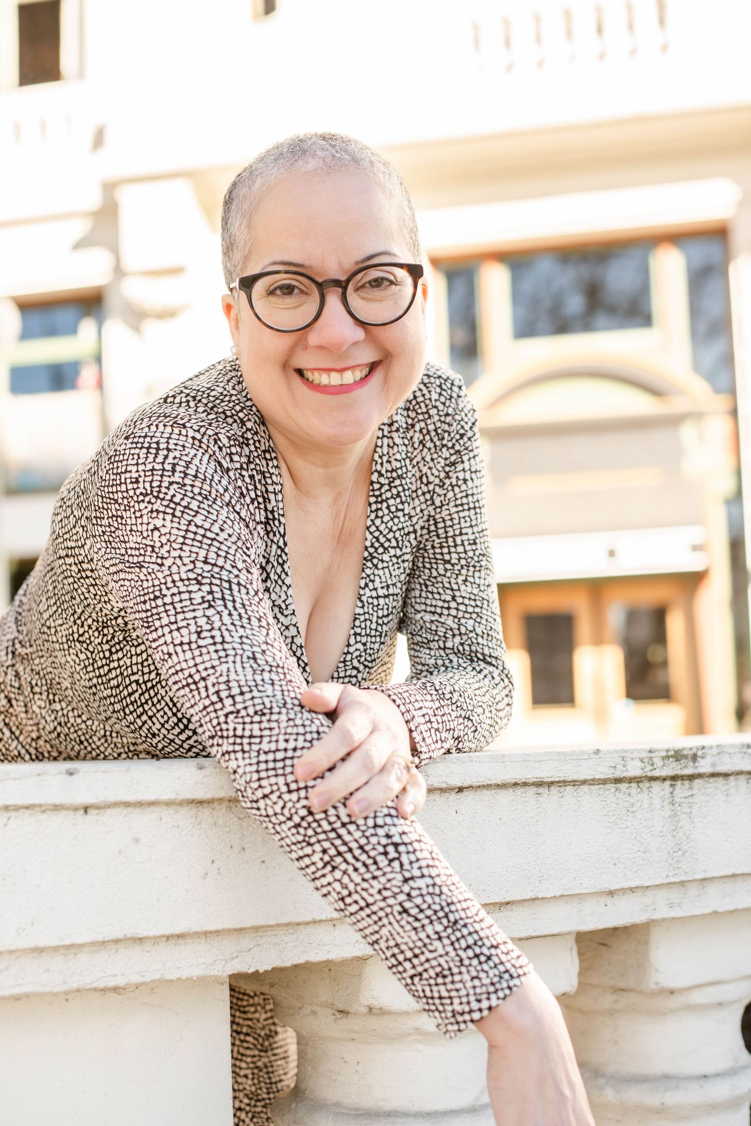 Professional headshots of someone in a patterned blouse leaning on concrete railing outdoors.