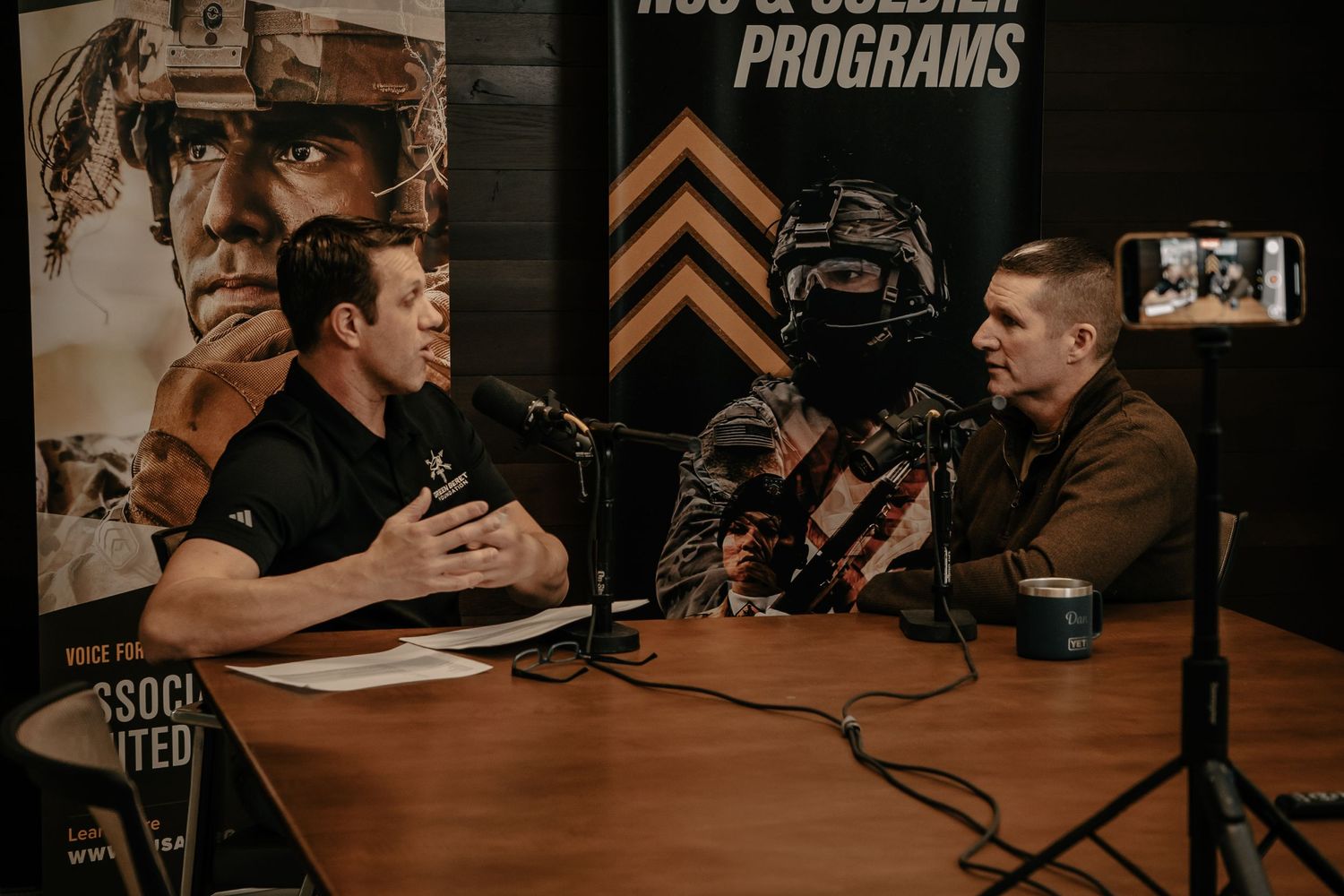 Two people engaged in conversation at a podcast recording setup with microphones and dark studio lighting.