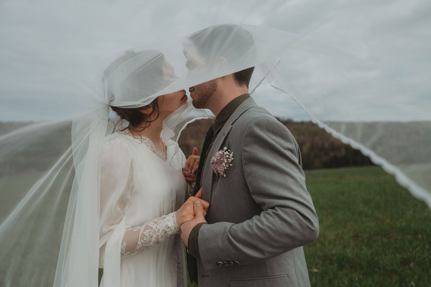 Intimate wedding portrait with windswept veil in cloudy countryside.